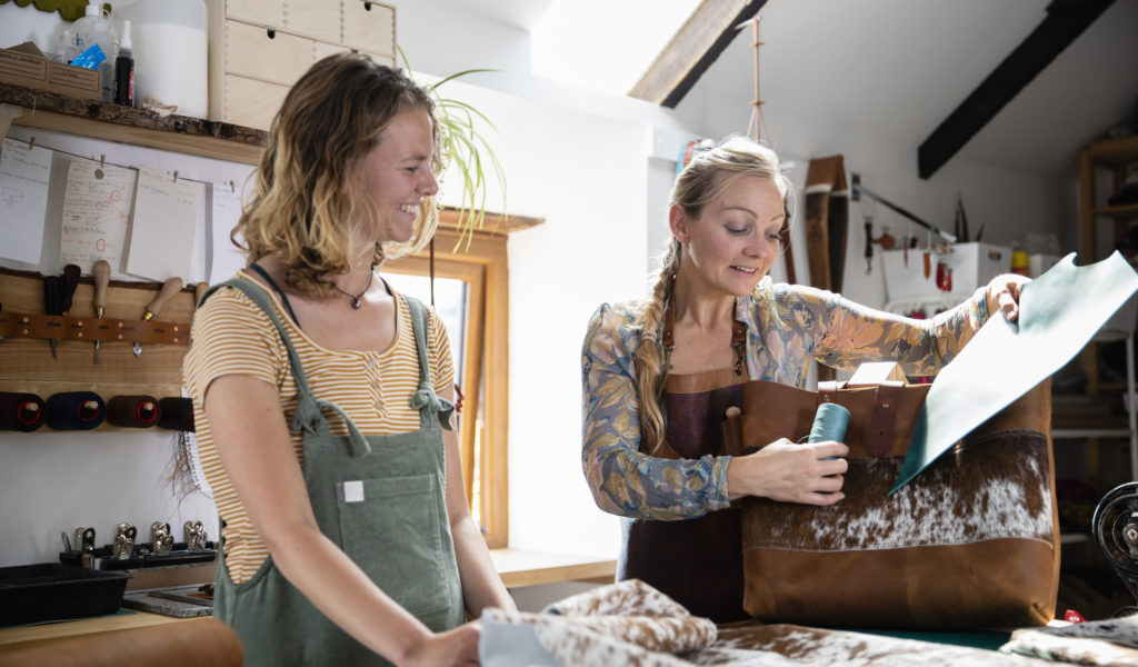 A tanner in her work studio, giving a leather product making workshop to one of her students. They are discussing a handbag that the teacher has made herself from leather, she is holding up blue material and blue thread against the bag, explaining how it could be customised further. The materials she uses are ethically sourced and she is committed to a zero-waste approach in her business. / Female Focus Collection